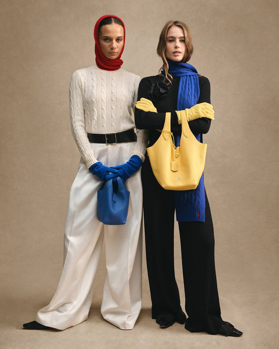 Two women standing, one in cream knit with red head covering and blue gloves, the other in black knit with a blue scarf and yellow bag.