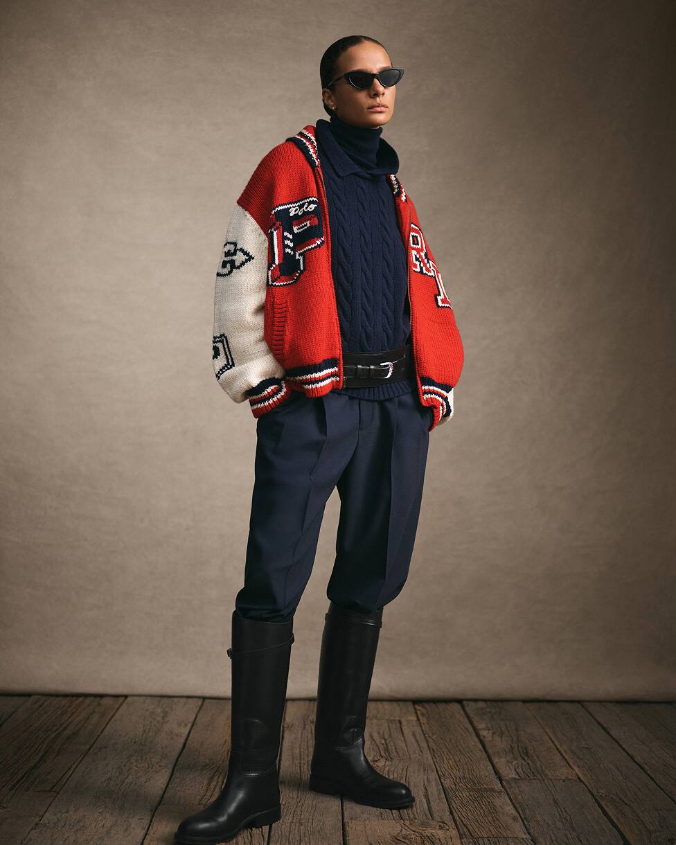 Woman standing wearing a red and white varsity jacket, navy jumper and trousers and tall black boots.